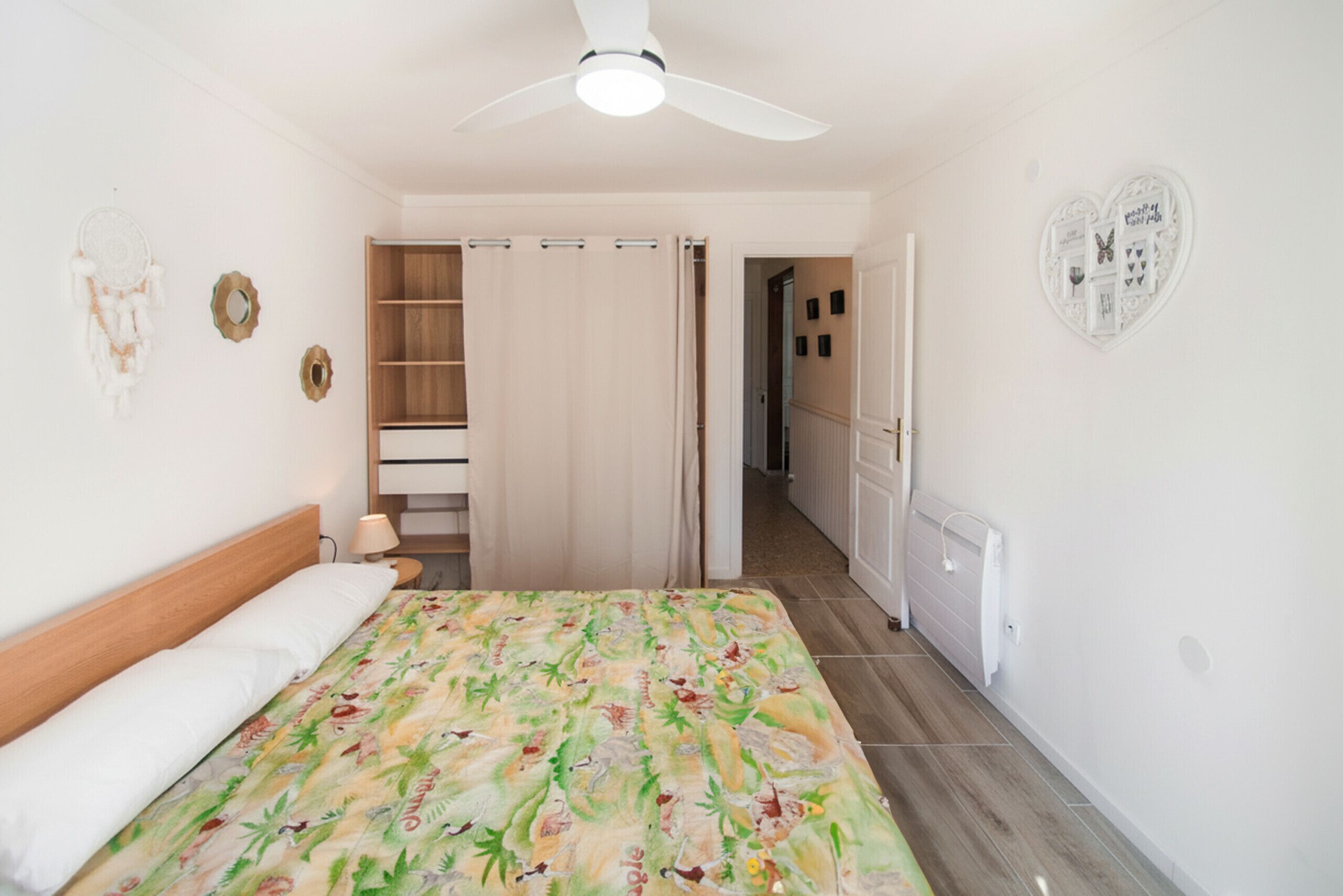 A bedroom with a bed, a ceiling fan, a wardrobe, and a hallway leading to a bathroom.