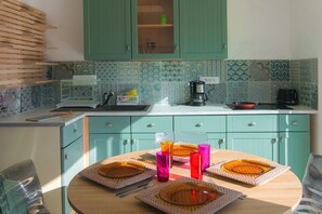 Eine Küche mit türkis-farbenen Schränken, einem runden Holztisch, der mit orangefarbenen Tellern, pinken Gläsern und einer Kaffeemaschine gedeckt ist.