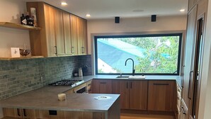 A modern kitchen with wooden cabinets, a large island with stools, a window with a view of greenery, and a black rug. - Modern Minimalist Cabin in quiet setting. 12-minute walk to reekside gondola. (Whistler)