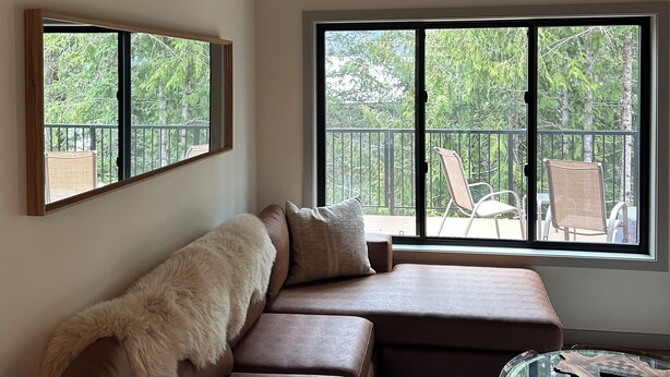 A modern living room with a brown leather sectional sofa, a glass coffee table, and a large window with a view of outdoor furniture. - Modern Minimalist Cabin in quiet setting. 12-minute walk to reekside gondola. (Whistler)