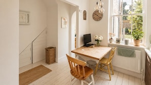 Une pièce lumineuse avec une table en bois, deux chaises, un ordinateur, une horloge et une fenêtre ornée de plantes.
