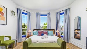 A bedroom with a bed, two bedside tables, a green chair, a green sofa, and a window with curtains. - Urban Haven Indy | Hot Tub + Game Room (Indianapolis)