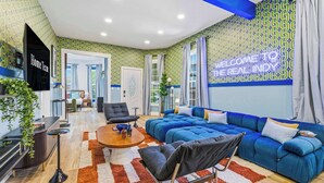A modern living room with a blue sofa, a coffee table, and a TV. There is a kitchen area in the background with a sign that reads 'Home Team.' - Urban Haven Indy | Hot Tub + Game Room (Indianapolis)