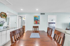 A dining area with a wooden table and chairs, a painting above the table, and a kitchen area with cabinets and a countertop. - Madeira Norte 406 - Premier (Madeira Beach)