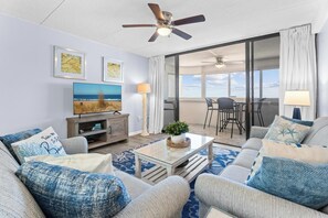 A living room with a sofa, coffee table, TV, and a balcony with a view. - Oceanfront Condo With Pool - Steps To The Beach (Ocean City)