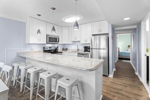 A modern kitchen with white cabinets, a central island with bar stools, stainless steel appliances, and pendant lighting. - Oceanfront Condo With Pool - Steps To The Beach (Ocean City)