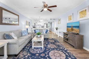 A living room with a sofa, coffee table, and TV, a kitchen in the background, and a ceiling fan. - Oceanfront Condo With Pool - Steps To The Beach (Ocean City)