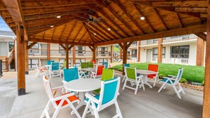 Eine Terrassensitzgelegenheit im Freien mit weißen Tischen und bunten Stühlen, darüber hölzerne Balken und im Hintergrund ein Gebäude.