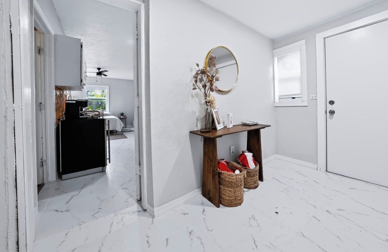 A hallway with a white door, a mirror, a small table with baskets, and a view into another room with a window.