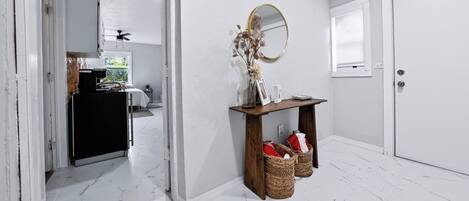 A hallway with a white door, a mirror, a small table with baskets, and a view into another room with a window.
