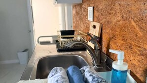 Una cocina con un lavamanos de acero inoxidable, una tabla de cortar de madera, un escurreplatos y una botella azul de jabón para manos.