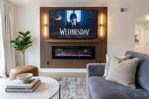 A living room with a flat-screen TV mounted on a wooden wall, a fireplace, a grey sofa, a round coffee table, and a plant.