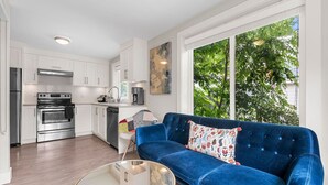 Et moderne køkken med stålemmer, et spiseområde med en blå sofa og et glasbord, samt et stort vindue med udsigt til træer.