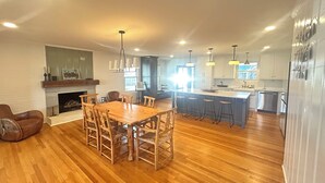 Amplia sala de estar con una mesa y sillas de madera para comer, una chimenea y una cocina con una isla grande y taburetes de bar.