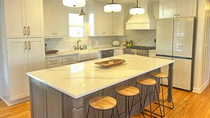 Una cocina moderna con armarios blancos, una isla grande con taburetes, iluminación colgante y un refrigerador de acero inoxidable.