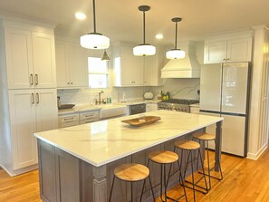 Una cocina moderna con armarios blancos, una isla grande con taburetes, iluminación colgante y un refrigerador de acero inoxidable.