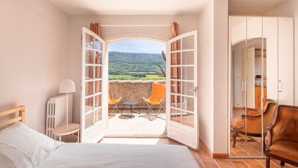 Ein Schlafzimmer mit offener Tür, die auf einen Balkon mit zwei Stühlen und einem Tisch führt. Man sieht ein Bett und einen Kleiderschrank.