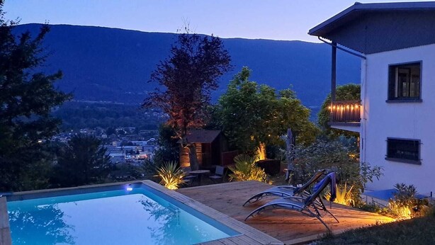 A swimming pool, lounge chairs, a building with a balcony, and a mountainous backdrop.