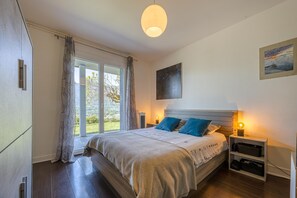 A bedroom with a bed, bedside tables, a nightstand, a window with curtains, and a painting on the wall.