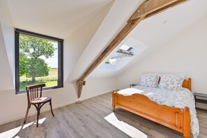 Ein Schlafzimmer mit schrägen Wänden, einem hölzernen Bett, einem Stuhl und einem Fenster mit Blick auf einen Baum.