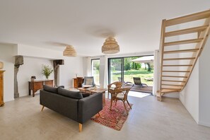 Ein modernes Wohnzimmer mit einer Couch, einem Couchtisch und einem Sessel. Rechts ist eine Treppe und durch große Fenster ist ein Blick ins Freie möglich.