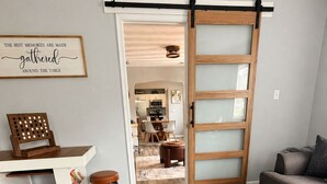 Un saló amb una taula baixa de fusta, un sofà, una zona de menjador amb una taula i cadires blanques, i una cuina al fons.