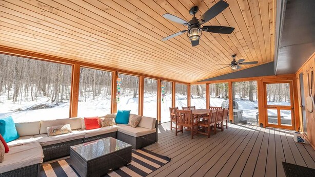 Eine Holzterrasse mit einer Couch, einem Couchtisch und einem Esstisch, große Fenster mit Blick auf Schnee und Deckenventilatoren.