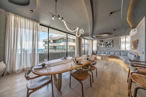 A modern dining area with a long wooden table, chairs, and a view of a building through large windows. - Oceanfront 3BR Luxury Maiim Tankah w/Pool & Gym (Tulum)