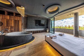 A modern bedroom with a large bed, a built-in bathtub, a flat-screen TV, and a large window with a view of greenery. - Oceanfront 3BR Luxury Maiim Tankah w/Pool & Gym (Tulum)