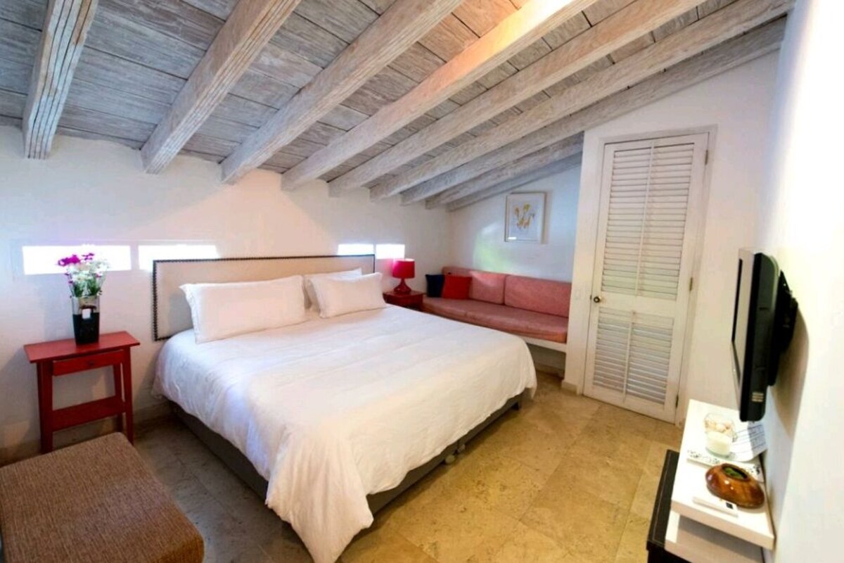 A bedroom with a large bed, a red side table, a red sofa, a white door, and a wooden ceiling.