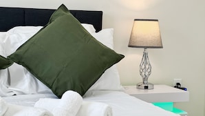 A neatly made bed with white linens, a green pillow, and rolled white towels. There is a lamp with a textured shade on a bedside table. - Ramblei near Drexel Park: 6-Bedroom Unit B (Philadelphia)