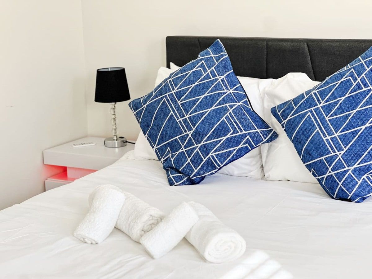 Een net opgemaakt bed met witte lakens en blauw gestreepte kussens, een witte nachtkastje met een lamp, en een opgerolde witte handdoek op het bed.