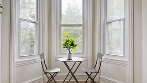 A corner room with three white-paneled windows, a round table with two chairs, and a potted plant with yellow flowers. - Ramblei near Drexel Park: 6-Bedroom Unit B (Philadelphia)