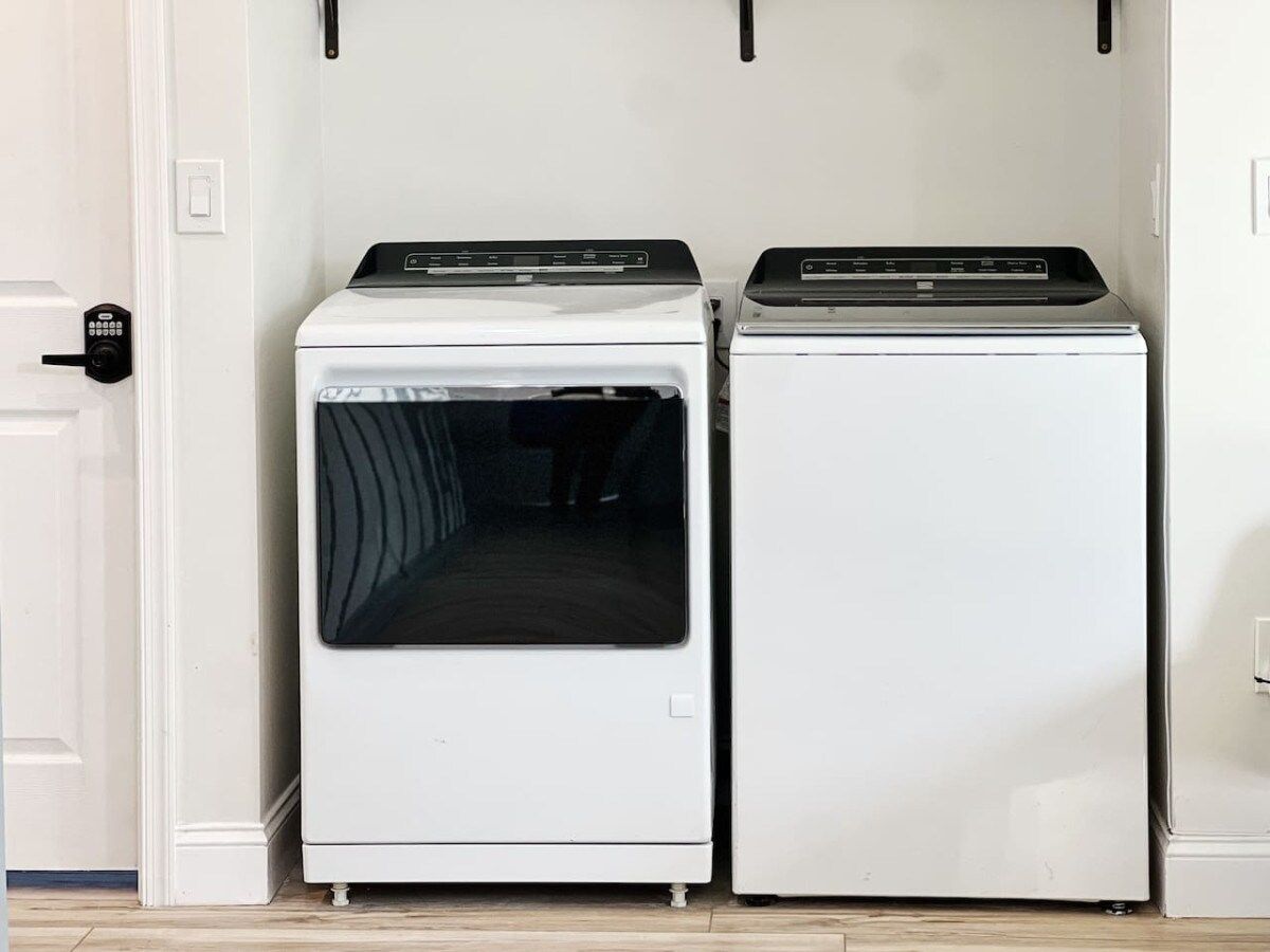 Two white appliances, likely a washer and dryer, in a room with a white door and a light switch on the wall.