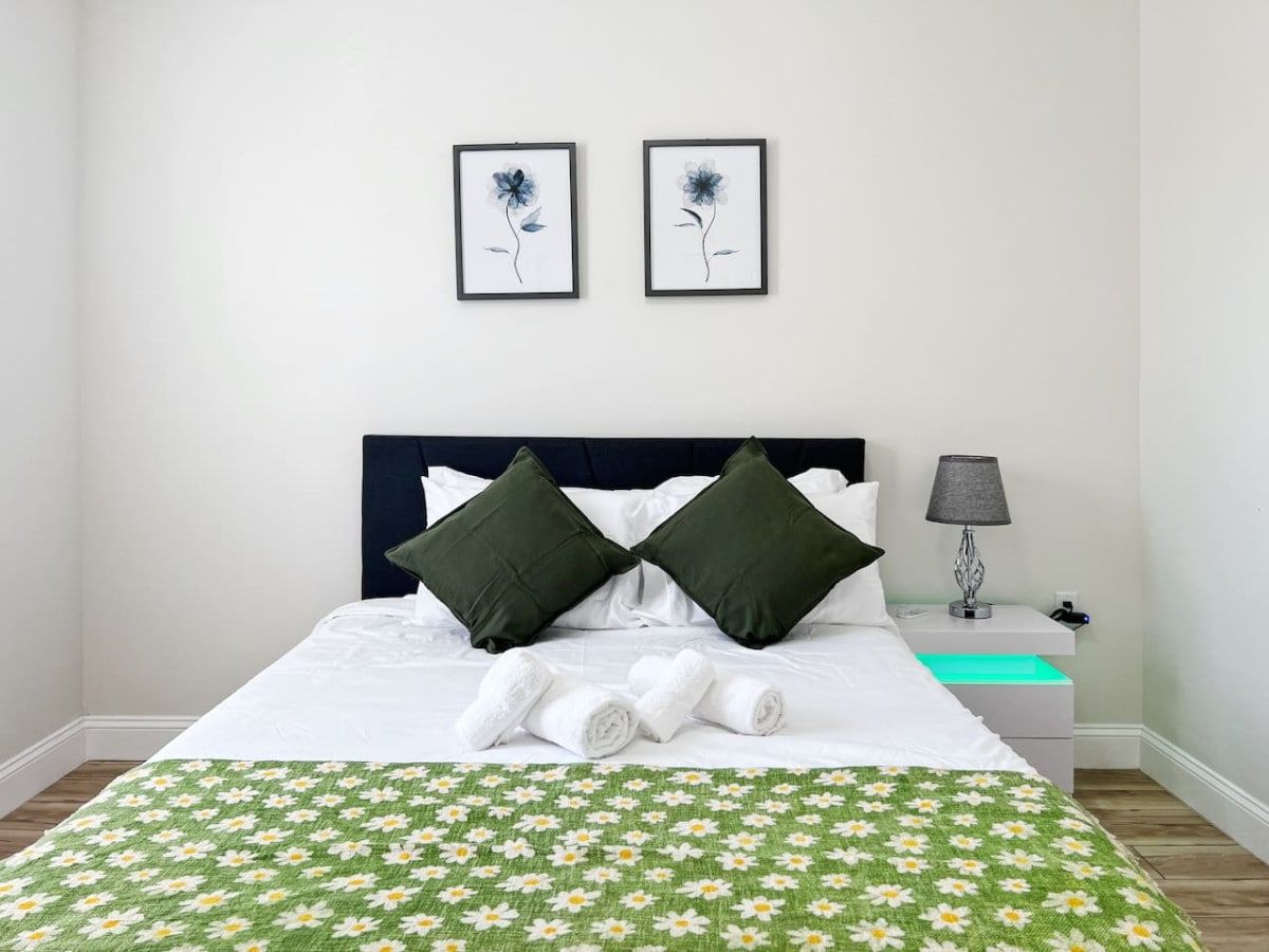 A neatly made bed with a green floral bedspread, two dark green pillows, and a framed picture above it.