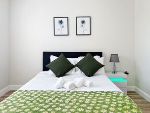 Een net opgemaakt bed met een groen bloemenbedover, twee donkergroene kussens en een lijst met een foto erboven.