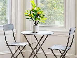 Une pièce avec une table blanche et deux chaises, une plante en pot posée sur la table, et une fenêtre donnant sur des arbres.