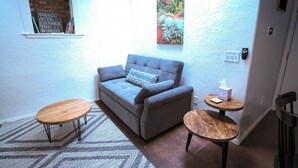 Ein gemütliches Wohnzimmer mit einem grauen Sofa, zwei Holztischen, einem gemusterten Teppich und einem an der Wand befestigten Regal mit Büchern und einem Schild.