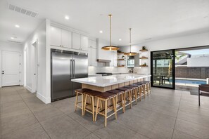 Eine moderne Küche mit einem Edelstahl-Kühlschrank, einer großen Kücheninsel mit Sitzgelegenheiten, Pendelleuchten und Blick auf einen Swimmingpool durch Glastüren.