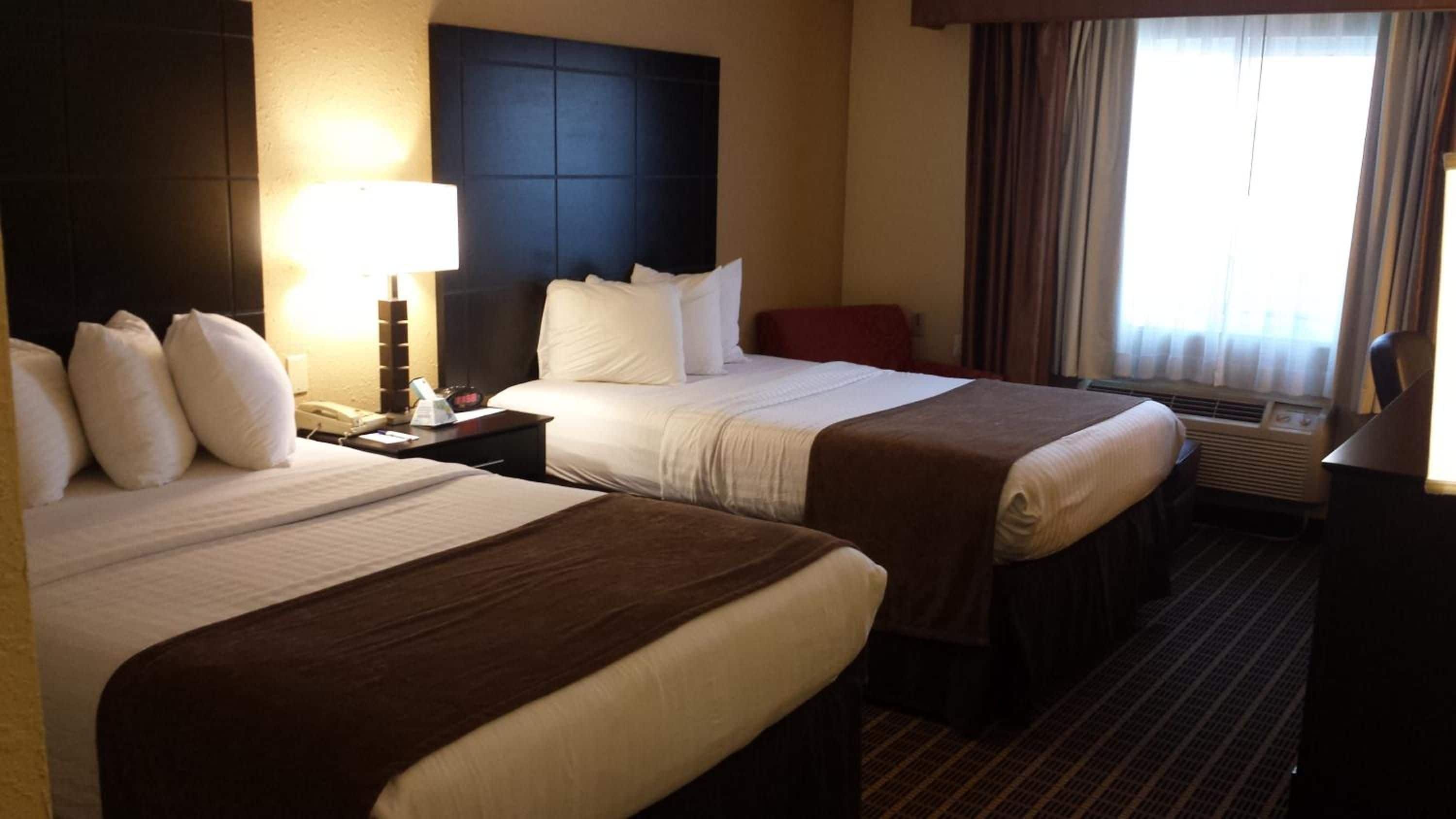 A hotel room with two beds, a bedside table with a lamp, a telephone, and a window with curtains.