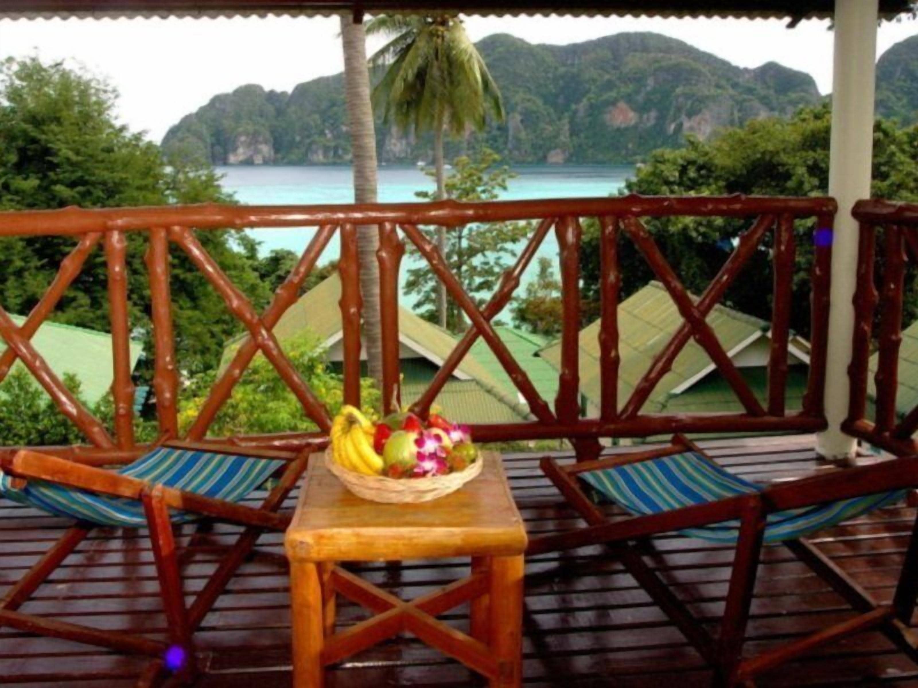 Une terrasse en bois avec deux chaises et une table, un panier en osier rempli de fruits, et une vue sur une mer turquoise et des collines verdoyantes.