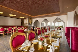 A restaurant with ornate ceiling, red velvet chairs, gold table settings, and a view of the outdoors. - Saura Hotel Agra A Club Mahindra (Agra)