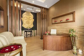 A reception area with a wooden reception desk, a sofa, a Buddha statue, and a wall-mounted shelf with flowers. - Saura Hotel Agra A Club Mahindra (Agra)