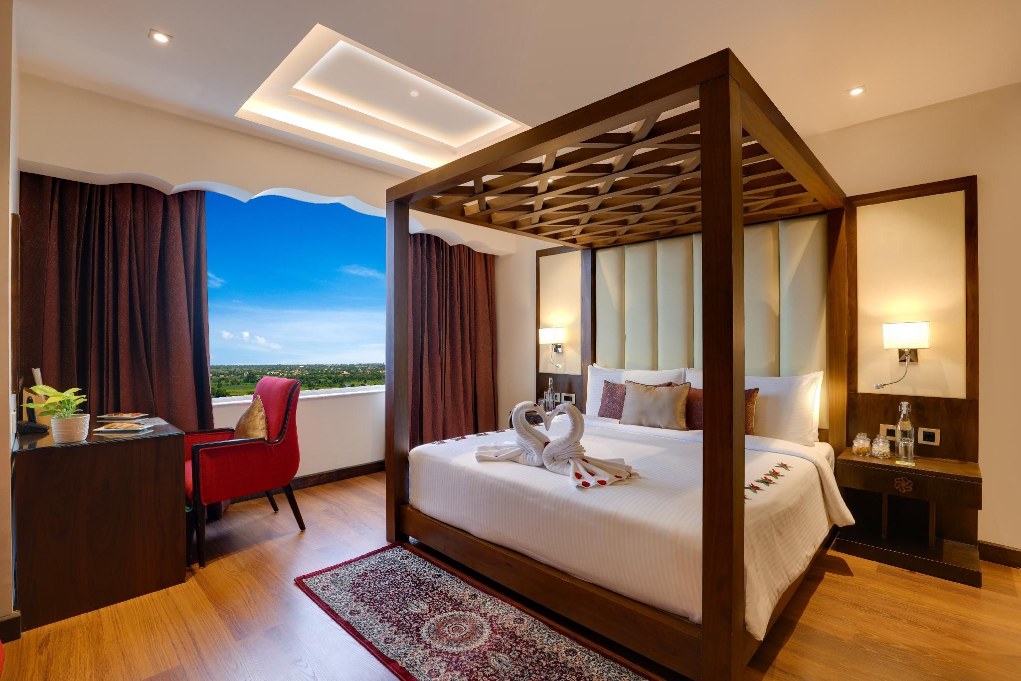 A hotel room with a four-poster bed, a desk with a chair, a bedside table, and a view of the landscape.