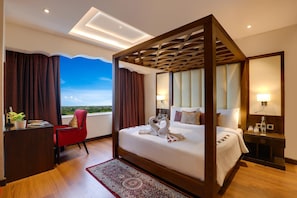 A hotel room with a four-poster bed, a desk with a chair, a bedside table, and a view of the landscape. - Saura Hotel Agra A Club Mahindra (Agra)