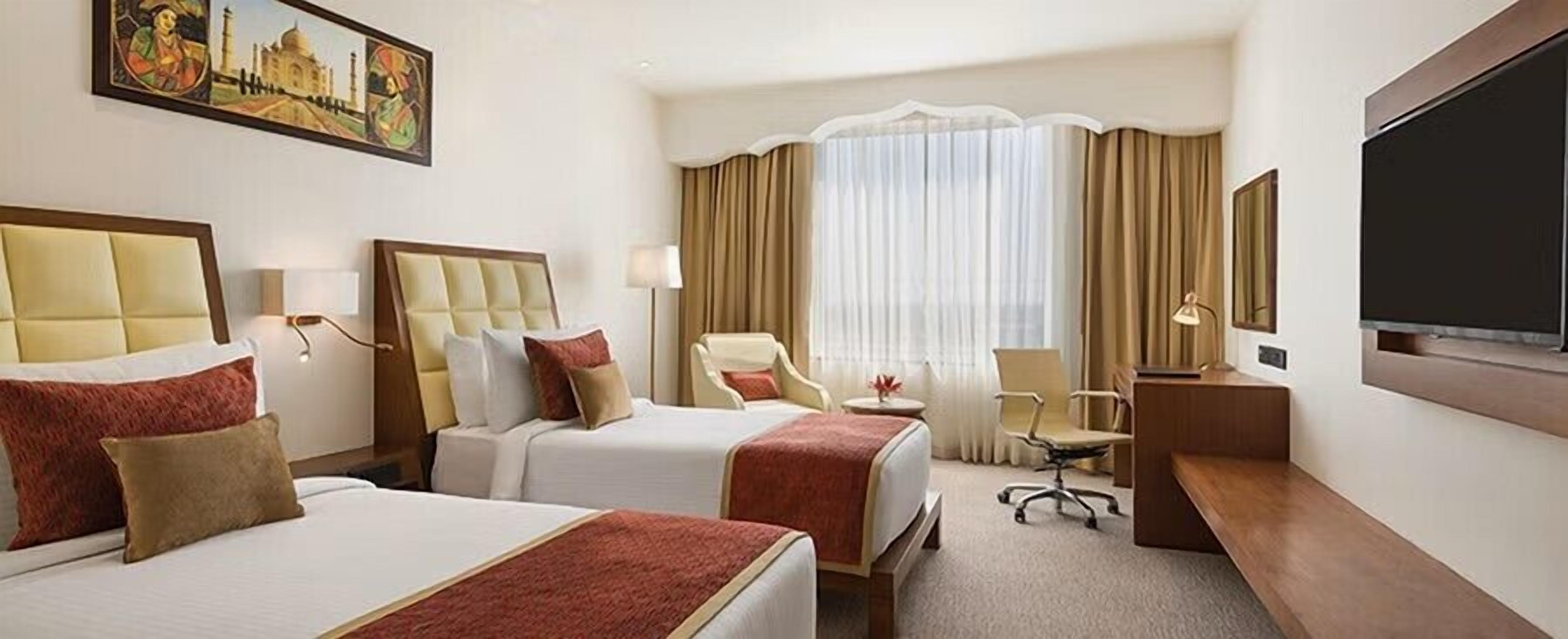A hotel room with two beds, a desk, a chair, a TV, and a large window with curtains.