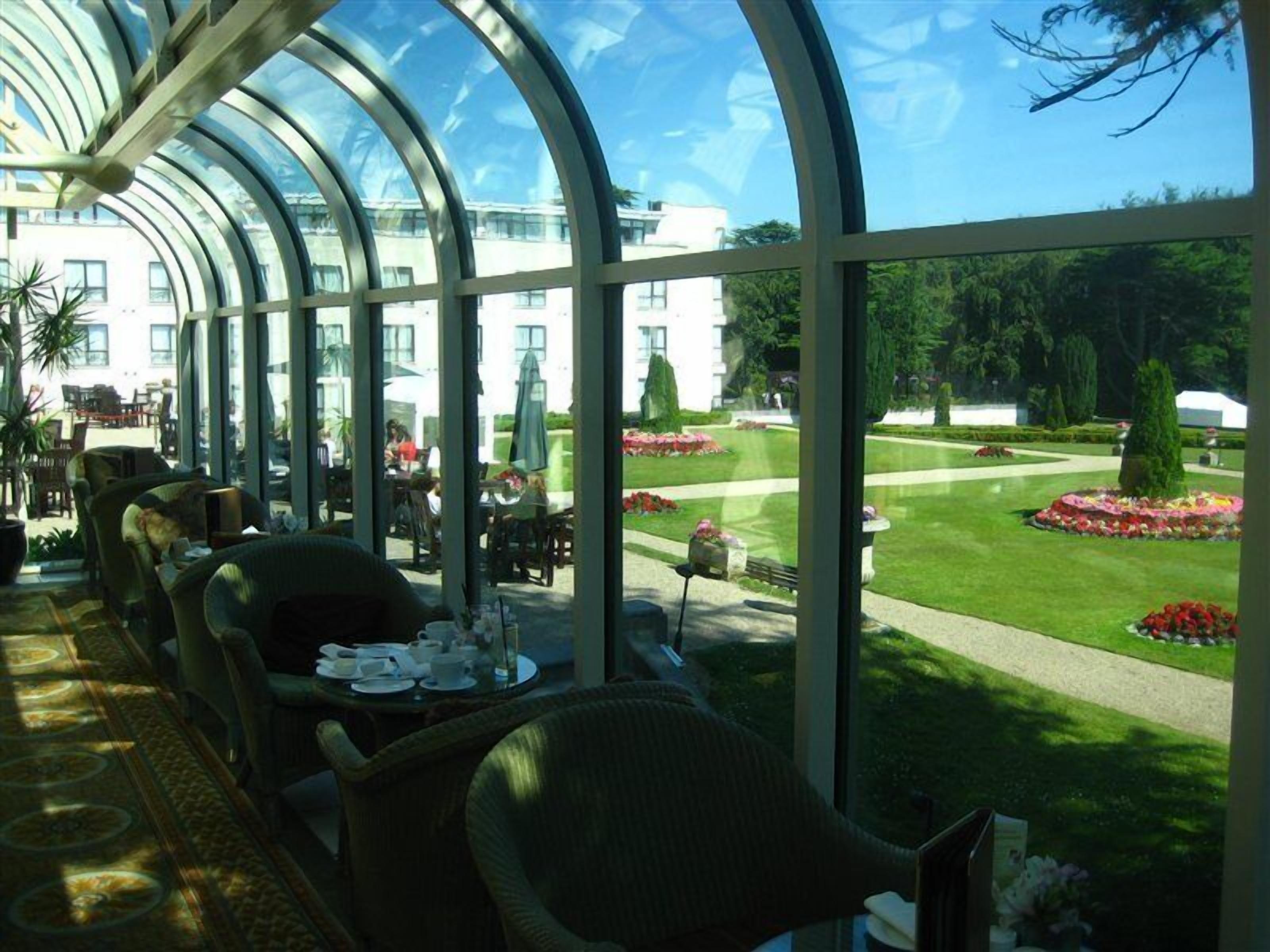 A conservatory with a glass ceiling, dining tables, chairs, and a view of a well-manicured garden.