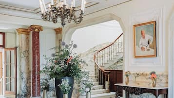 A grand foyer with a checkered floor, a large chandelier, marble columns, a staircase, and a painting on the wall.