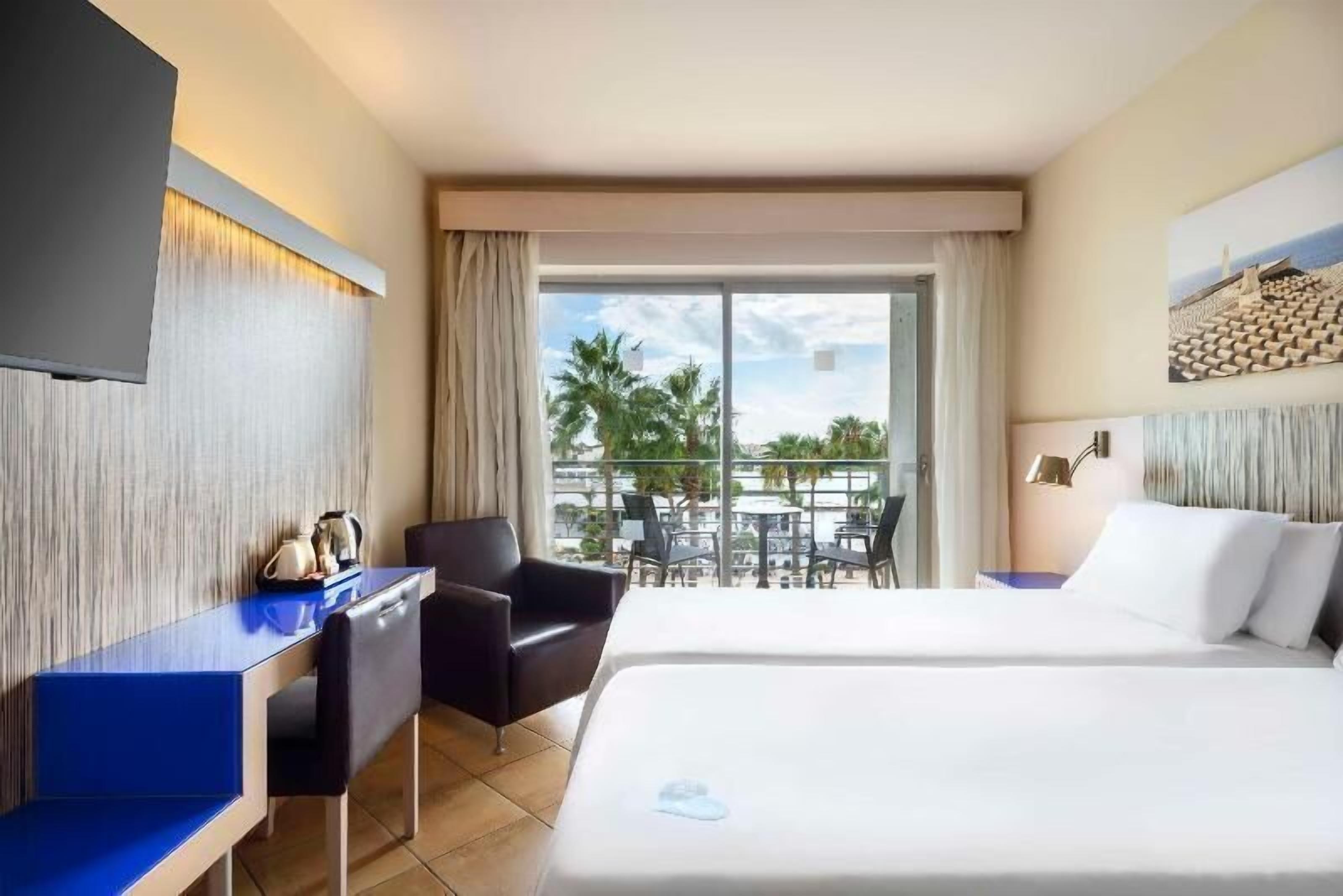 A hotel room with a bed, a desk, a chair, a TV, and a balcony with outdoor seating.