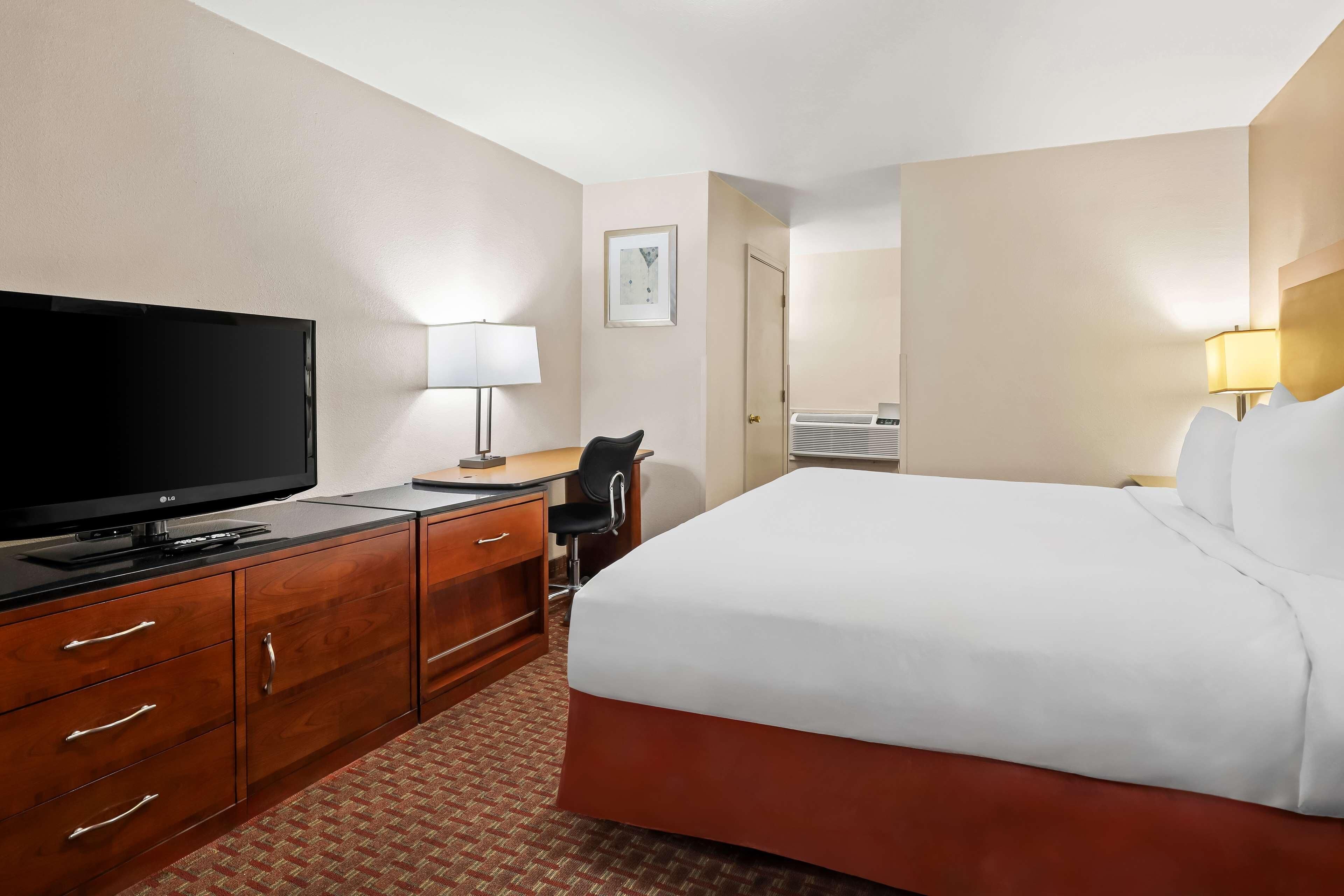 A hotel room with a bed, a desk with a TV, a chair, a lamp, and a door.